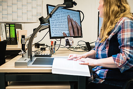 Eine weiblich gelesene Person mit blonden langen Haaren und Sonnenbrille auf dem Kopf sitzt an einem Bildschirm mit großer Schrift. Sie nutzt unter anderem eine Kamera um Texte aus Büchern auf einem Bildschirm größer lesen zu können.  