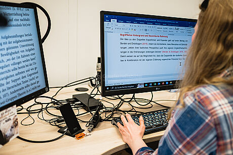 Eine weiblich gelesene Person mit blonden langen Haaren und Sonnenbrille auf dem Kopf sitzt an einem Bildschirm mit großer Schrift. Sie nutzt unter anderem eine Kamera um Texte aus Büchern auf einem Bildschirm größer lesen zu können.  